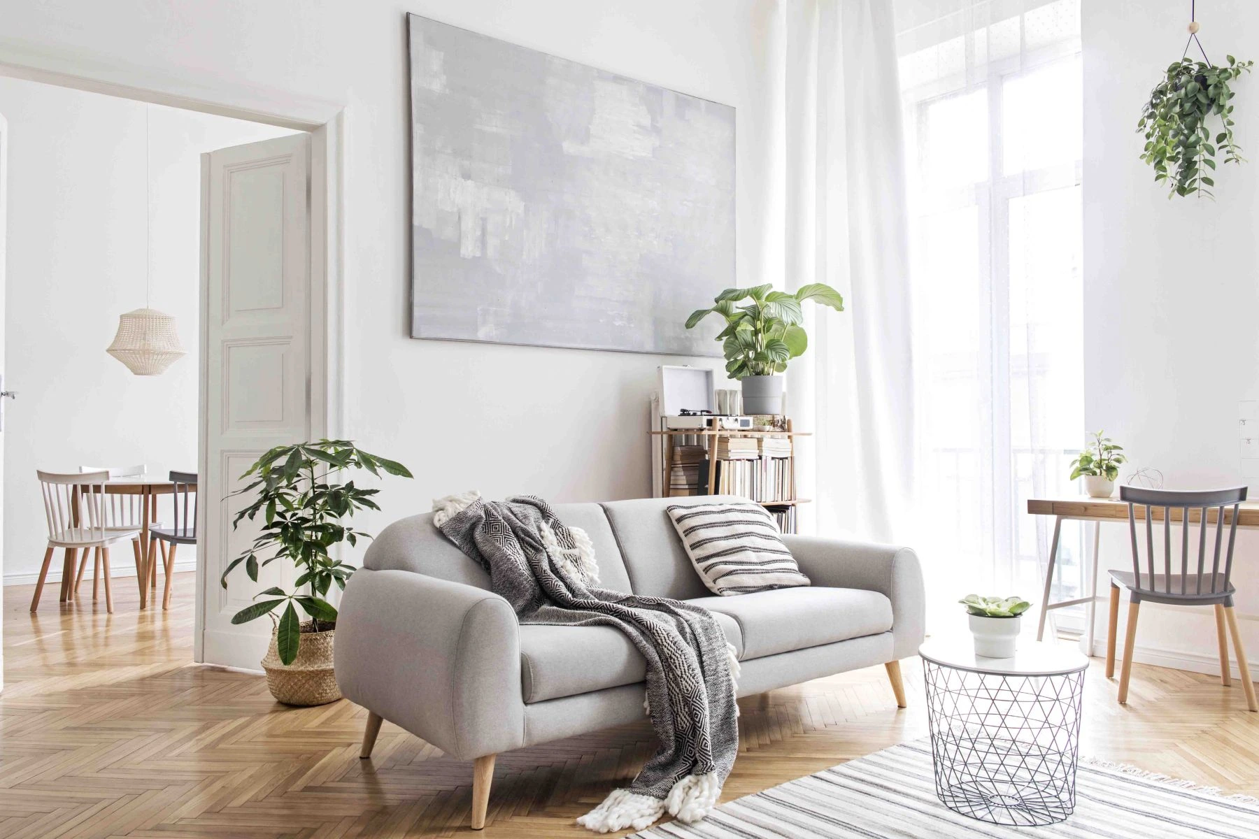 Stilvolles skandinavisches Wohnzimmer mit Designermöbeln, Pflanzen, Bambus-Bücherständer und Holztisch. Braunes Holzparkett. Abstraktes Gemälde an der weißen Wand.